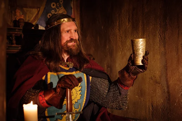 Old medieval king with goblet of wine on the throne in ancient castle interior. — Stock Photo, Image