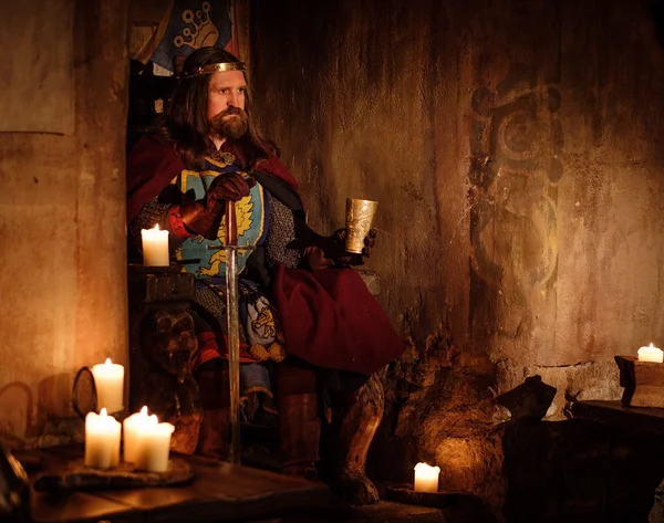 Old medieval king with goblet of wine on the throne in ancient castle interior. — Stock Photo, Image