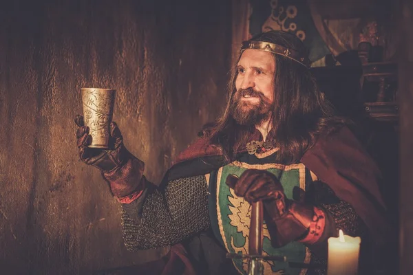 Old medieval king with goblet of wine on the throne in ancient castle interior. — Stock Photo, Image