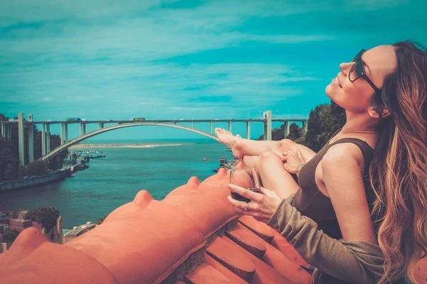 Frau genießt Sonnenbad mit einem Glas Portwein auf dem Dach, auf der Flussbrücke Hintergrund. — Stockfoto