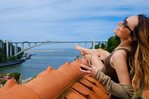 Жінка насолоджується сонячною ванною з келихом портового вина на даху, на фоні річкового мосту . — стокове фото