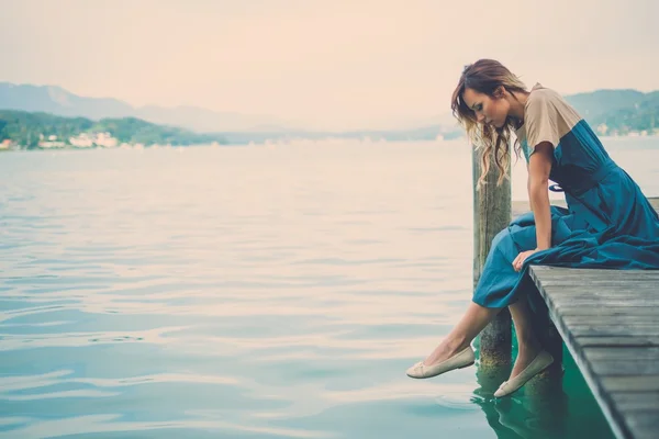 Välklädd kvinna som sitter på träpiren med floden bergsutsikt. — Stockfoto
