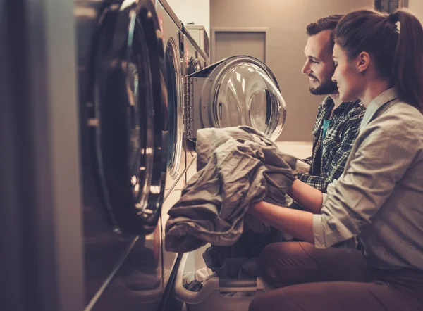 Joven pareja alegre haciendo la colada juntos en la lavandería . — Foto de Stock