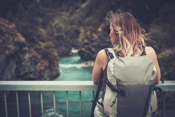 Mulher caminhante com mochila em pé na ponte sobre um rio de montanha selvagem . — Fotografia de Stock