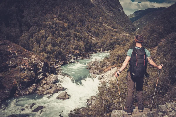 Man hiker with backpack standing on the edge of the cliff with epic wild mountain river view. — Stock Photo, Image
