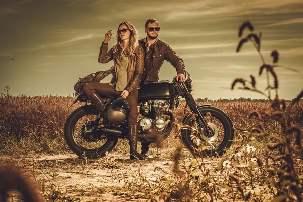Stijlvol café racer koppel op de vintage aangepaste motorfietsen in een veld. — Stockfoto