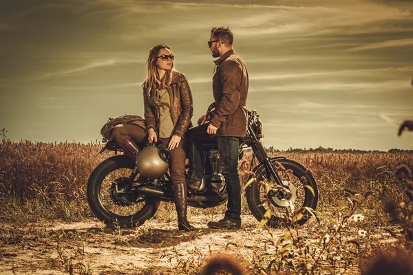 Casal de piloto de café elegante nas motocicletas personalizadas vintage em um campo . — Fotografia de Stock