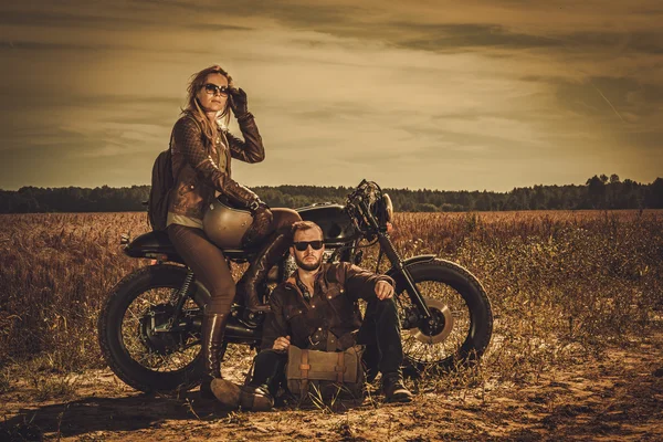 Casal de piloto de café elegante nas motocicletas personalizadas vintage em um campo . — Fotografia de Stock