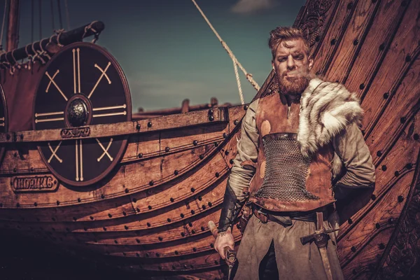 Viking krijger met assen permanent in de buurt van Drakkar aan de kust. — Stockfoto