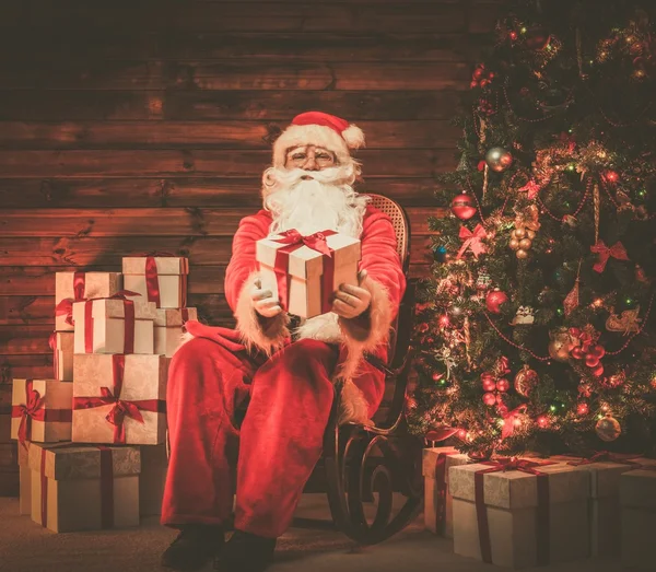 Papai Noel sentado na cadeira de balanço em casa de madeira interior apresentando caixa de presente — Fotografia de Stock