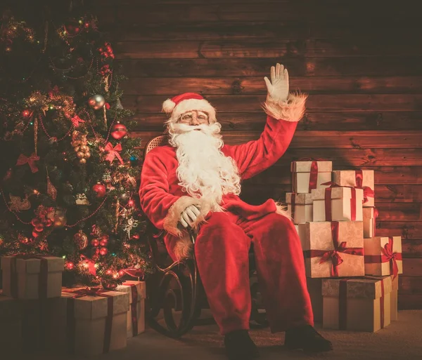 Papai Noel sentado em cadeira de balanço em interior de casa de madeira com caixas de presente ao seu redor — Fotografia de Stock