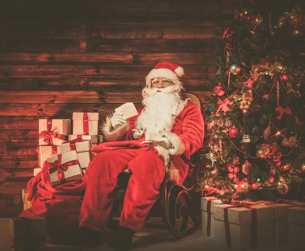 Père Noël assis sur chaise à bascule dans l'intérieur de la maison en bois avec des lettres dans les mains — Photo