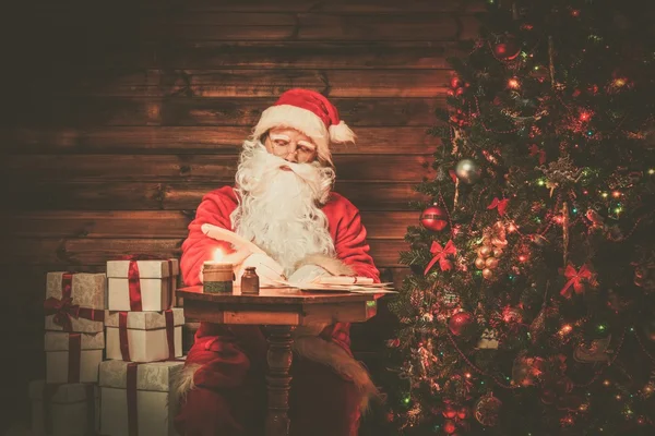 Santa Claus in wooden home interior sitting behind table and writing letters with quill pen — Stock Photo, Image