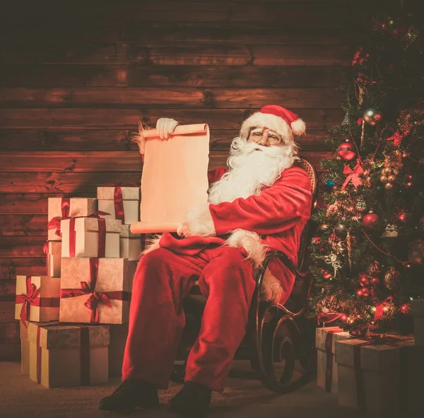 Papai Noel em casa de madeira interior segurando rolagem lista de desejos em branco — Fotografia de Stock