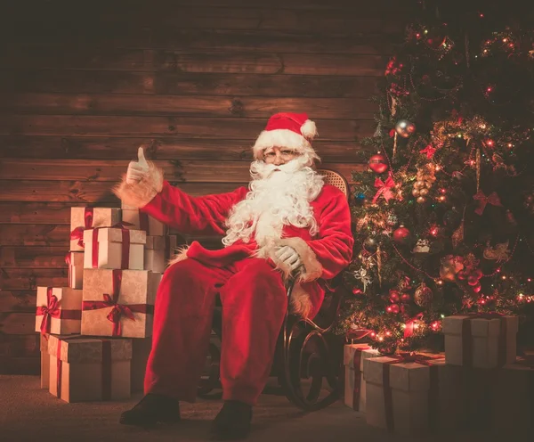 Santa Claus sitting on rocking chair in wooden home interior with gift boxes around him — Stock Photo, Image