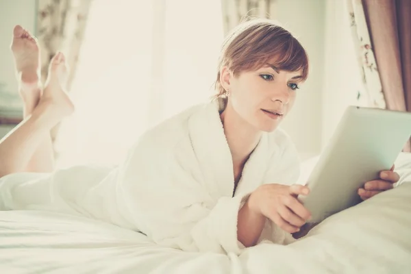 Schöne Frau im Bademantel auf einem Bett mit Tablet-PC liegend — Stockfoto