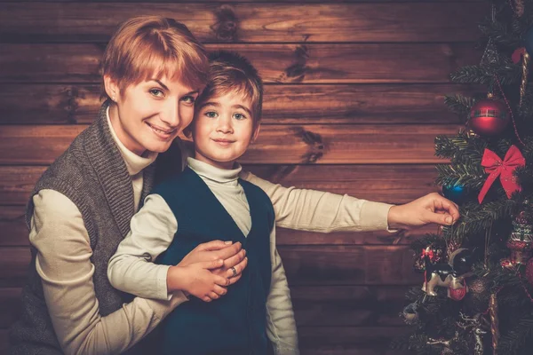 Glückliche Mutter und ihr kleiner Junge schmücken Weihnachtsbaum im Holzhaus-Interieur — Stockfoto