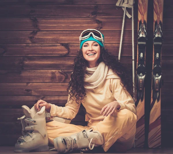 Happy woman with skis and ski boots sitting near wooden wall in snowflakes — Stock Photo, Image