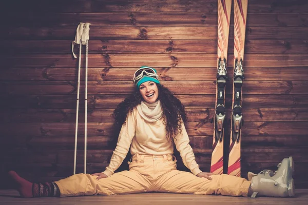 Happy woman with skis and ski boots sitting near wooden wall in snowflakes — Stock Photo, Image