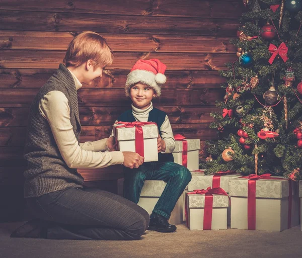 Mother and lIttle boy with gift box under christmas tree in wooden house interior — Stock Photo, Image