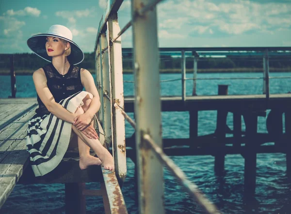 Mulher bonita usando chapéu e cachecol branco sentado no velho cais de madeira — Fotografia de Stock