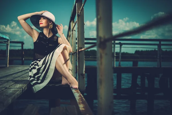 Beautiful woman wearing hat and white scarf sitting on old wooden pier — Stock Photo, Image