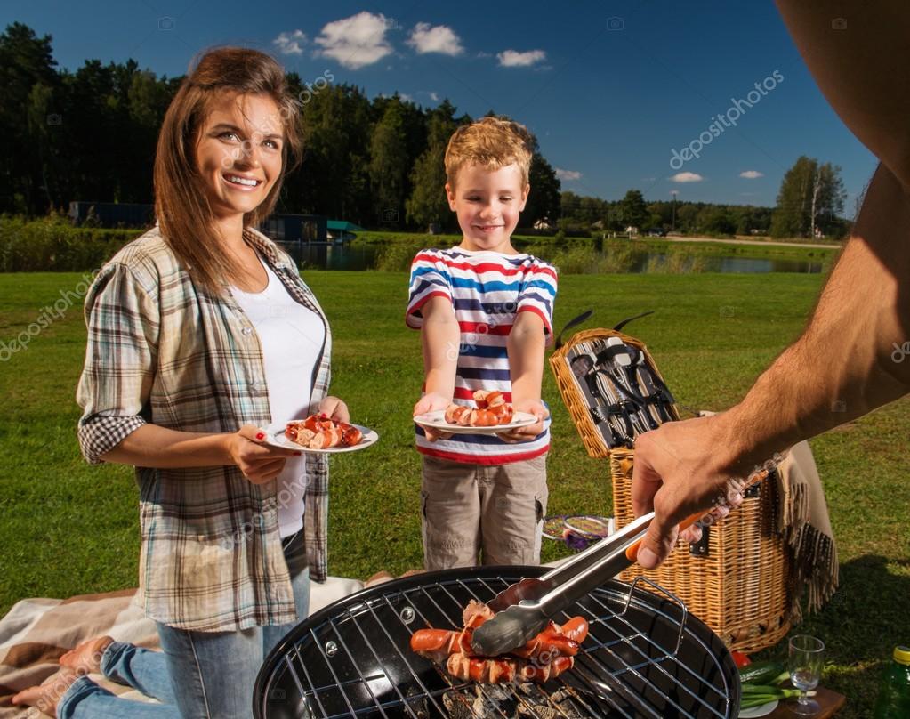 Young family preparing sausages on a grill outdoors — Stock Photo