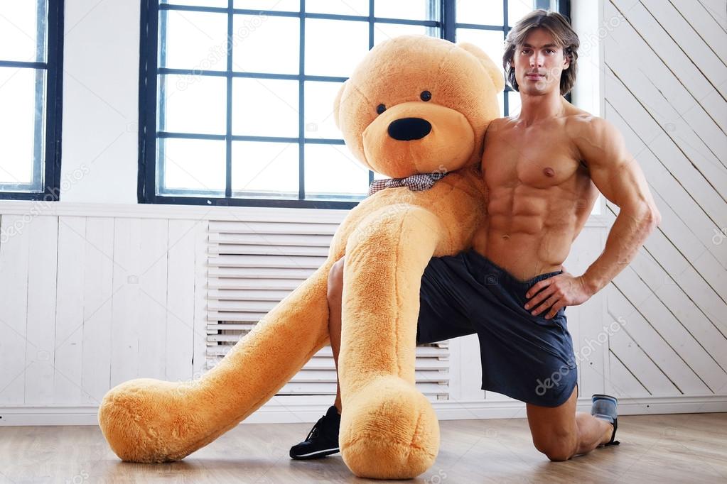 Download - Shirtless sporty male posing with big teddy bear on a floor. 