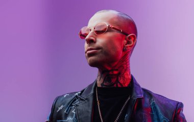 Deri ceketli dövmeli adam parlak arkaplanda poz veriyor.