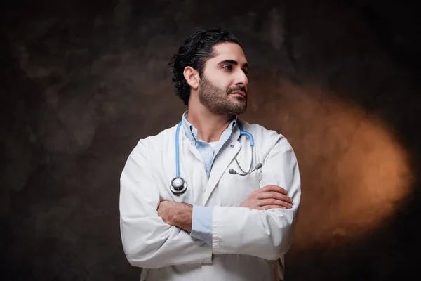 Mature and skilled doctor in labcoat poses in dark background — Stock Photo, Image