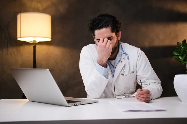 Doutor cansado no casaco senta-se à mesa com laptop em fundo escuro — Fotografia de Stock