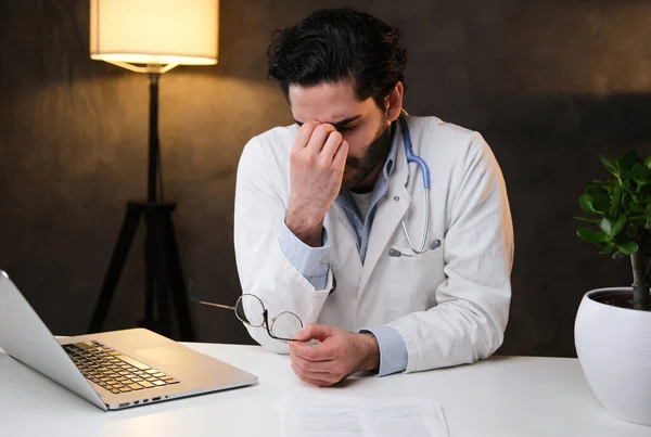 Уставший доктор в пальто сидит за столом с ноутбуком на темном фоне — стоковое фото
