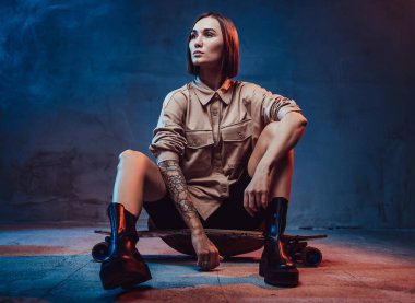 Dövmeli ve kısa saçlı neşeli kız yerde kaykayla oturuyor.