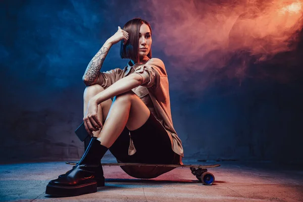 Hipster girl with tattoo and phone sits on skate in smokey background — Stock Photo, Image