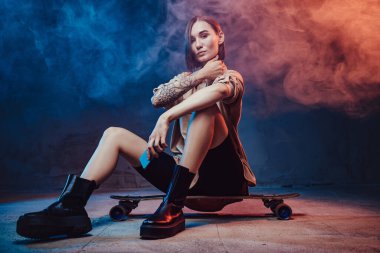 Dövmeli ve telefonlu hippi kız arka planda kaykay yapıyor.