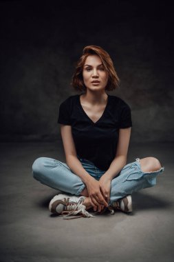 Siyah gömlekli kadın, siyah arka planda, yerde oturuyor.