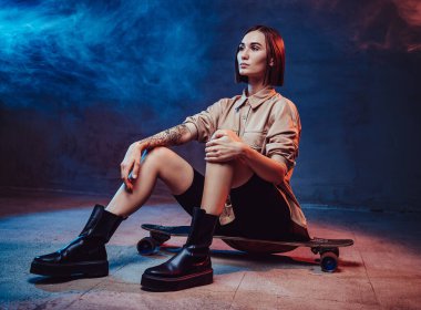 Güzel ve sportif bir kız. Saç kesimi ve sağ elinde dövmesi var. Kaykayın üstünde oturuyor..