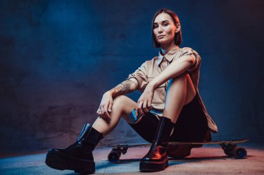 Dövmeli, kısa saçlı, çekici ve şık bir kadın kaykayın üzerinde oturuyor ve akıllı telefonunu tutuyor..