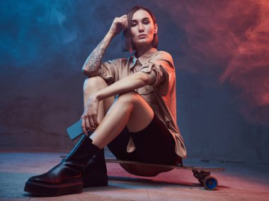 Dövmeli ve kısa saçlı sportif ve şık bir kadın kaykayın üzerinde oturuyor. Akıllı telefonunu dumanlı arka planda tutuyor..