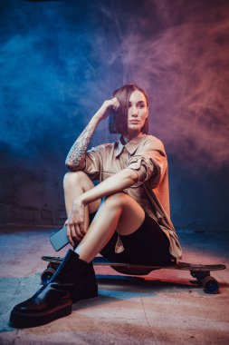 Kahverengi saçlı, dövmeli ve akıllı telefonlu güzel kız kaykayın üzerinde otururken dumanlı arka planda poz veriyor..