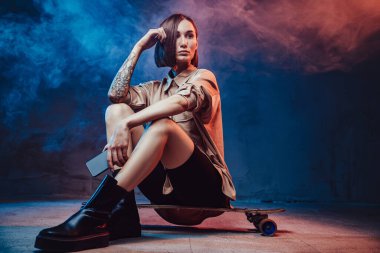 Dövmeli ve kısa saçlı sportif ve şık bir kadın kaykayın üzerinde oturuyor. Akıllı telefonunu dumanlı arka planda tutuyor..