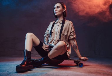 Dövmeli, kısa saçlı, şık giyinmiş çekici genç bir kadın model, arka planda dumanlı bir kamerayla paten üzerinde oturuyor..