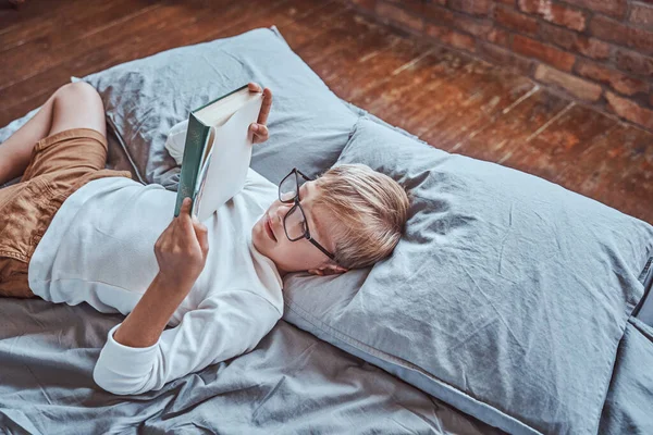 Chico positivo leyendo un libro en la sala de estar acostado en una cama — Foto de Stock