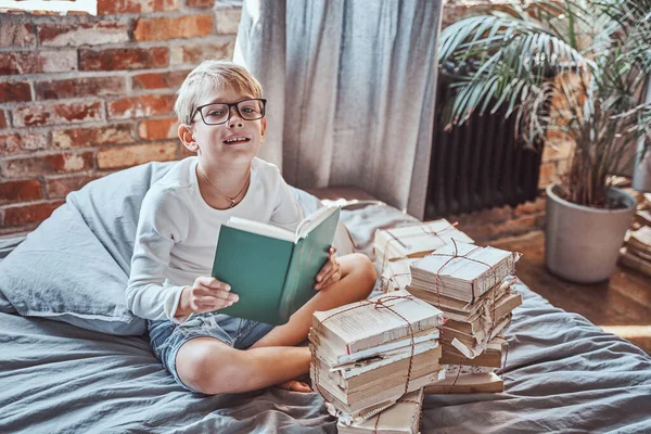 Niño inteligente con gafas lee libro sentado en una cama en el apartamento — Foto de Stock