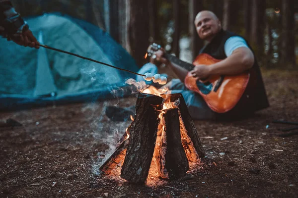 Marshmallow toasting on bonfire and bald man playing guitar
