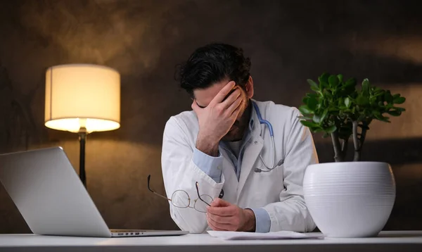 Unavený doktor sedí u stolu s rukou na čele v pokoji — Stock fotografie