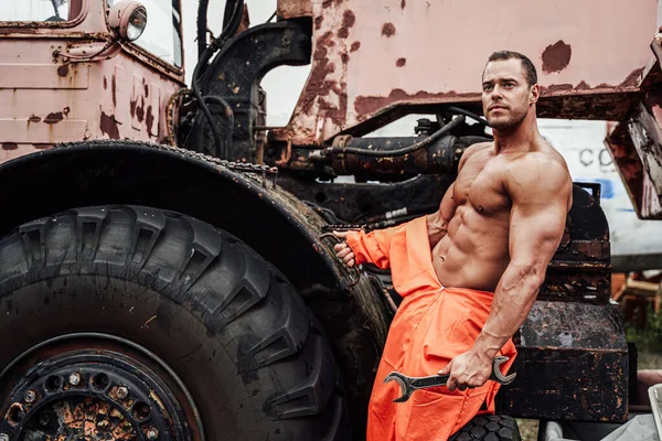 Handsome bodybuilder in orange uniform near abandoned vehicle — Stock Photo, Image