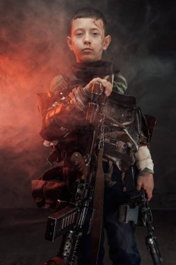 Dumanlı arka planda tüfeği olan küçük vahiysel çocuk portresi.