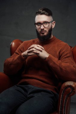 Koyu arka planda deri koltukta oturan dalgın sakallı kişi.