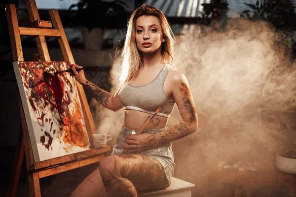 Mooie vrouw met tattoos schilderend op doek in de kamer — Stockfoto